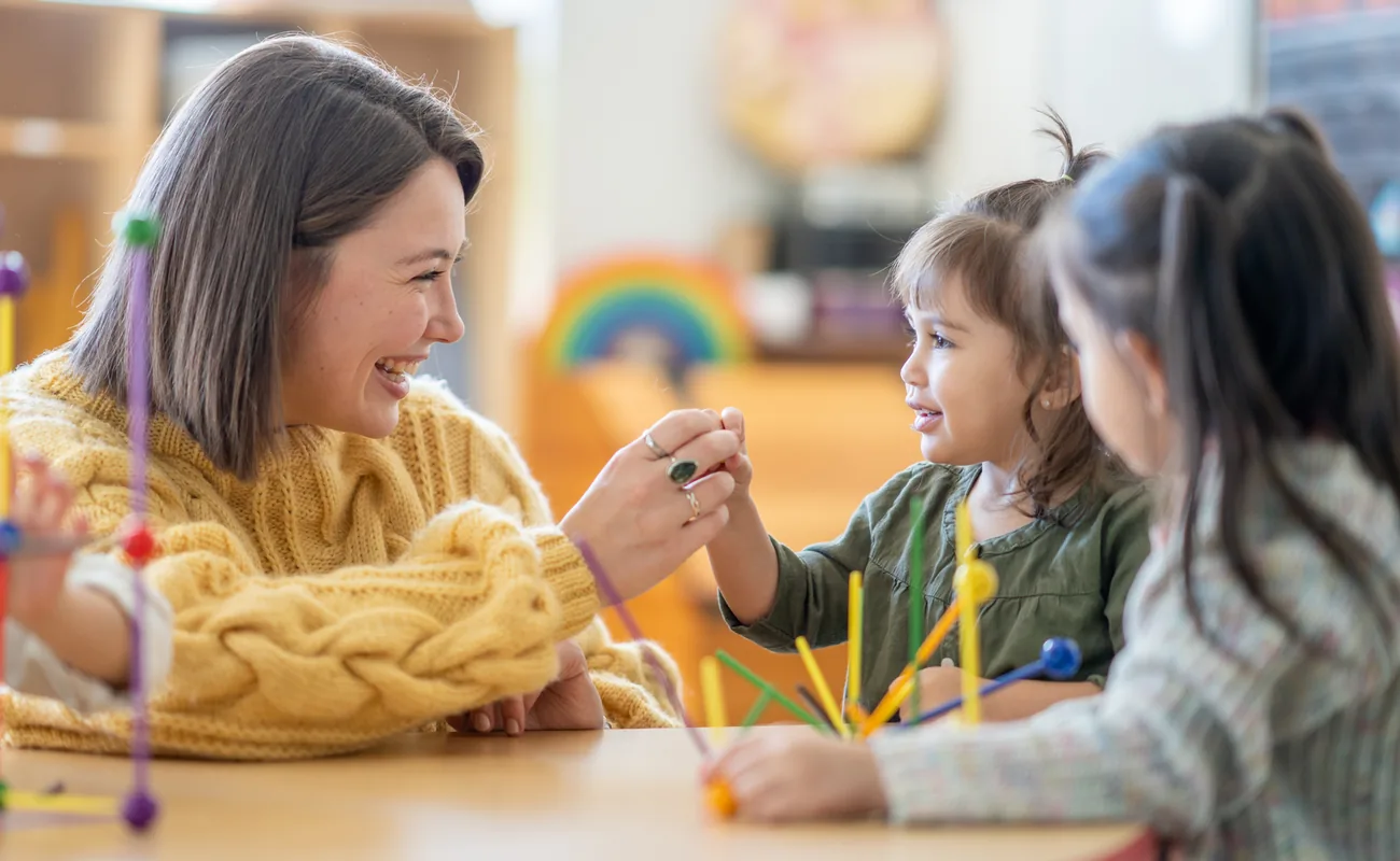 A smiling educator and a toddler building with colourful pieces at a table