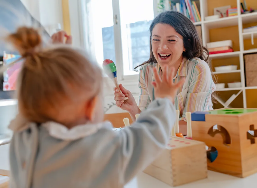 A toddler and a smiling educator playing with colourful maracas